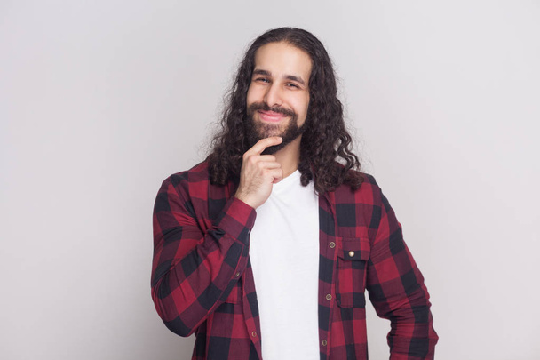 Handsome man with beard and black long curly hair in checkered red shirt touching chin and looking at camera while dreaming on grey background - Foto, immagini