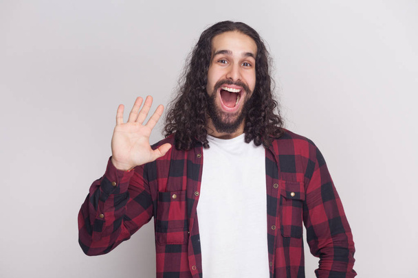 Happy surprised man with beard and black long curly hair in checkered red shirt looking at camera and greeting on grey background - Фото, изображение