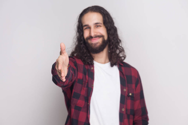 Happy smiley handsome man with beard and black long curly hair in checkered red shirt giving handshake and greeting with smile on grey background - Foto, Imagen