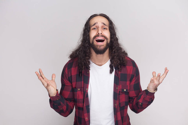 sad unhappy man with beard and black long curly hair in checkered red shirt standing with raised arms and looking at camera while asking on grey background - Photo, Image