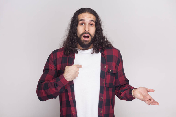 surprised handsome man with beard and black long curly hair in checkered red shirt looking at camera and pointing himself on grey background - Photo, Image