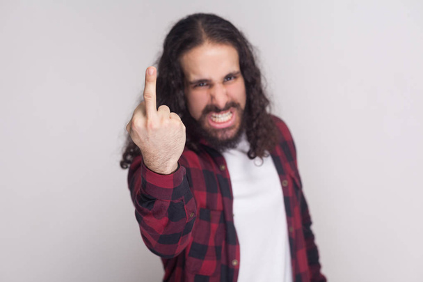 Portrait of angry handsome man with beard and black long curly hair in checkered red shirt looking at camera and showing middle finger on grey background - Φωτογραφία, εικόνα