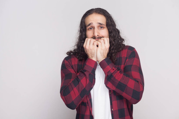 Portrait of nervous man with beard and black long curly hair in checkered red shirt looking at camera and biting nails on grey background - Фото, изображение