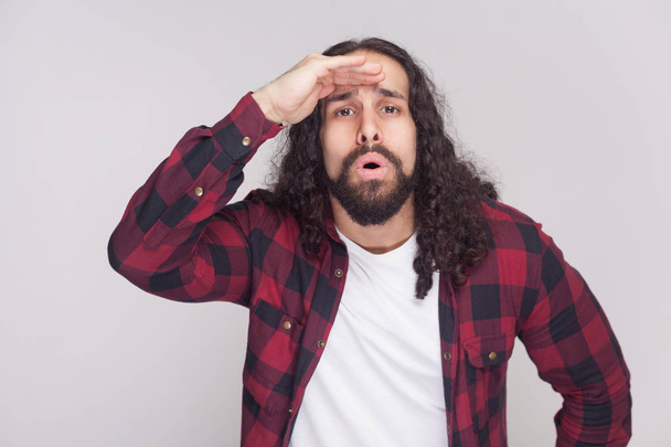 Portrait of surprised man handsome man with beard and black long curly hair in checkered red shirt standing and looking far with amazed face on grey background - Φωτογραφία, εικόνα