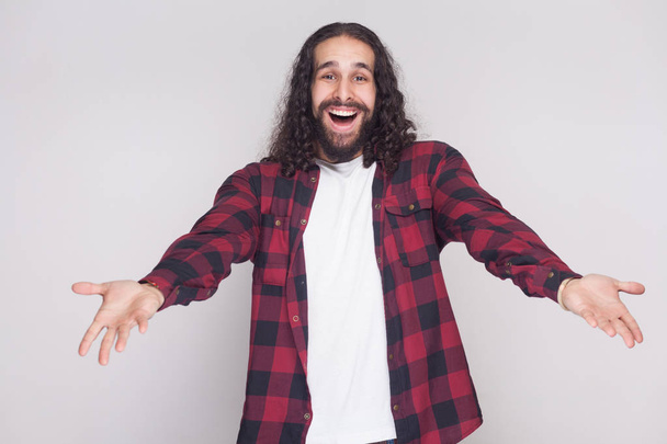 Surprised man with beard and black long curly hair in checkered red shirt looking at camera with raised arms on grey background - Zdjęcie, obraz