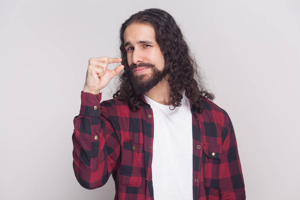 handsome man with beard and black long curly hair in checkered red shirt standing and pleading few more time while showing little sign on grey background - Foto, immagini