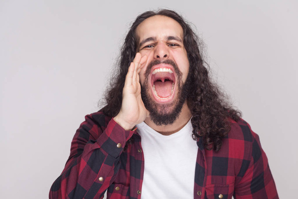 Portrait of handsome man with beard and black long curly hair in checkered red shirt standing and screaming with big open mouth on grey background - Photo, image