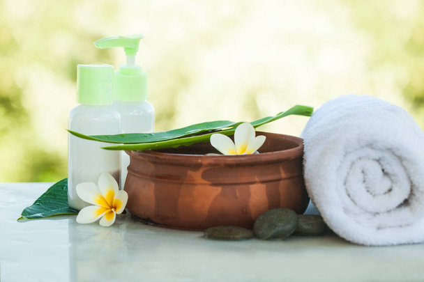 Spa or wellness setting outdoors with tropical flowers, bowl of water, towel and cream tube with sun light. Body care and spa concept - Photo, Image