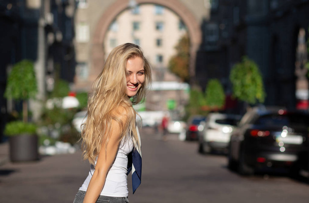Hymyilevä blondi malli pitkät hiukset yllään huivi ja valkoinen paita poseeraa auringon valossa kadulla
 - Valokuva, kuva