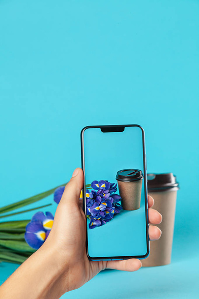 Takeaway paper coffee cup mockup isolated on blue background. Concept photo of coffee cup with smartphone and flowers. Free empty blank copy space for your design text or banner of brand. Trending colors - Photo, Image