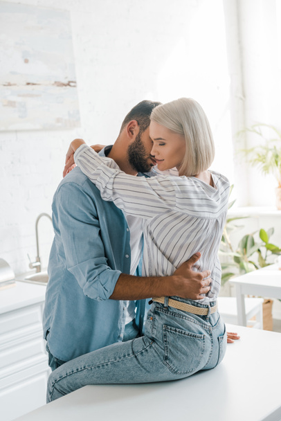 affectionate young couple hugging in kitchen, girlfriend sitting on kitchen counter - Fotografie, Obrázek