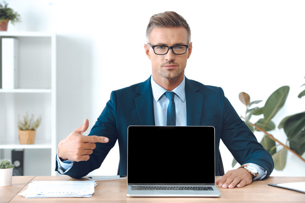 businessman pointing at laptop with blank screen and looking at camera - Foto, imagen