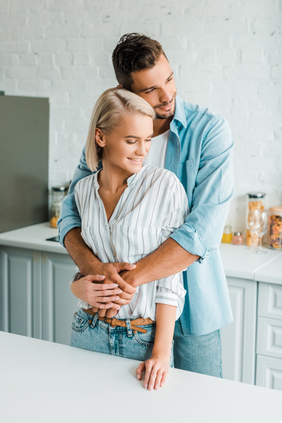 sensual smiling boyfriend hugging girlfriend from back in kitchen - Fotó, kép