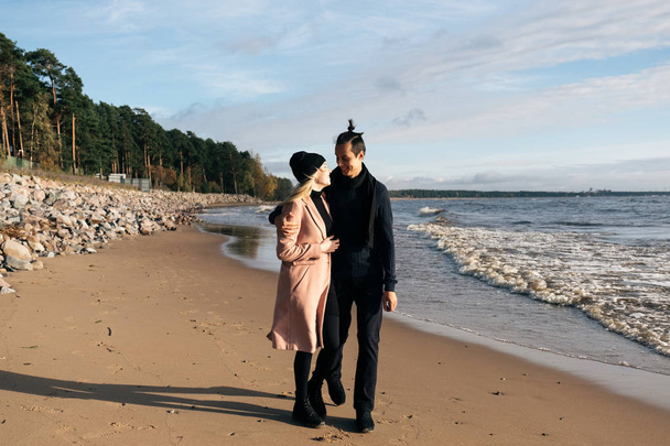 Young sensual couple having fun walking and hugging on beach. Cold autumn weather, coastline, park and ocean on the background. - 写真・画像