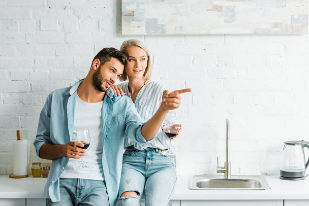 boyfriend holding glass of wine and pointing on something to girlfriend in kitchen - Photo, Image