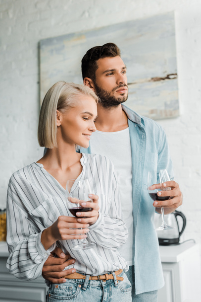 young couple hugging, holding glasses of red wine and looking away in kitchen - 写真・画像