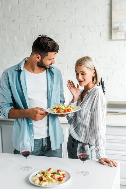 surprised girlfriend gesturing and looking at plate of salad in kitchen - Zdjęcie, obraz