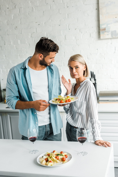 irritated girlfriend grimacing and gesturing near boyfriend with plate of salad in kitchen - Foto, Bild