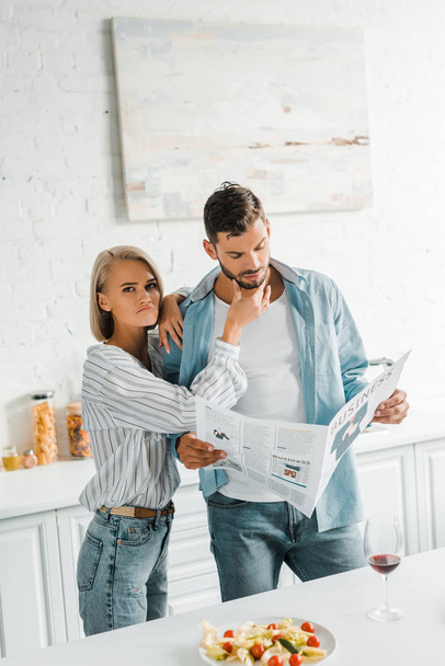 boyfriend reading newspaper and grimacing girlfriend touching his chin in kitchen - Photo, Image