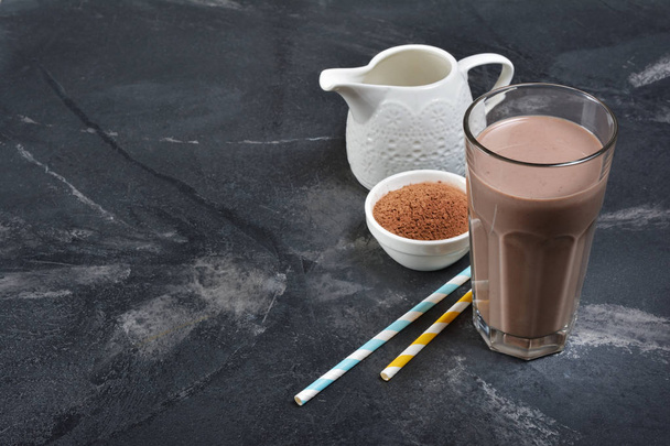 Glass of delicious cold chocolate milk drink with white jug and cocoa powder on black marble background - Zdjęcie, obraz