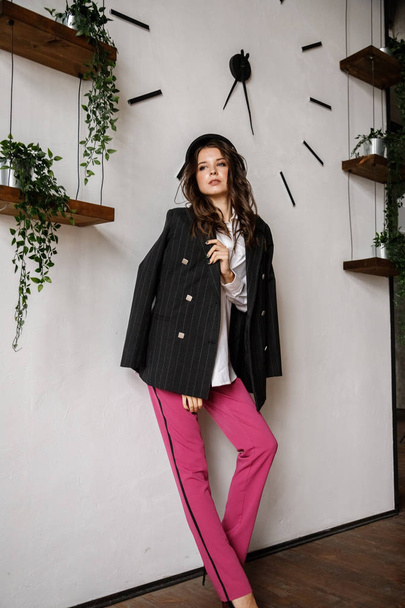 Модный портрет модели брюнетки в черной куртке, белой рубашке и розовых брюках. Молодая красивая женщина позирует в роскошных апартаментах интерьера
 - Фото, изображение
