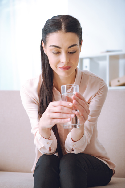 attractive smiling young woman sitting on couch and holding glass of water - Foto, afbeelding