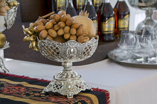 Frutti di legno decorativi in vaso d'argento tradizionale su arazzo armeno con bottiglie di alcol e bicchieri da vino su sfondo sfocato
 - Foto, immagini