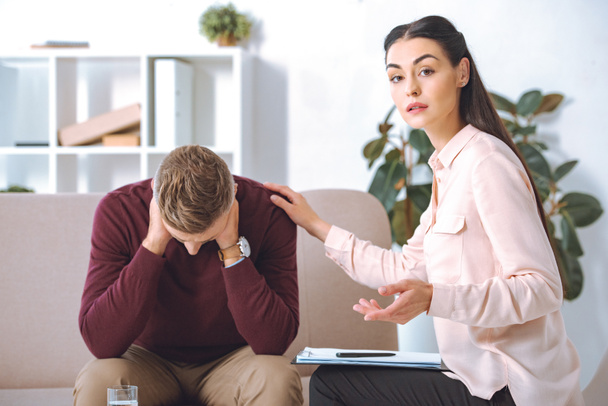 psychotherapist looking at camera while supporting upset patient in office - Fotoğraf, Görsel