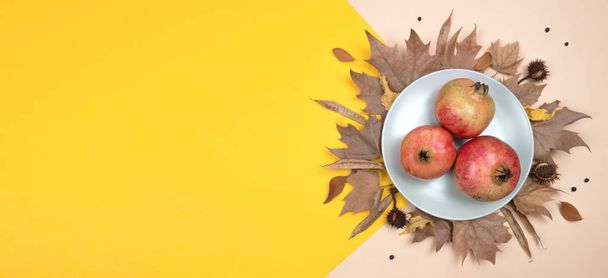 Creative Autumn layout. Plate with pomegranates on dried leaves, chestnuts and other elements on yellow and ocher background. Flat lay top view. Copy space. - Photo, Image
