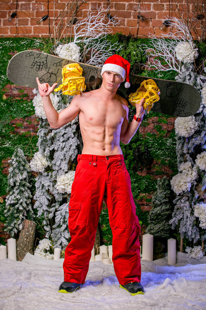young sexy sporty young man in red pants and Santa's cap keeps snowboard on his shoulders, vertical view - Фото, зображення