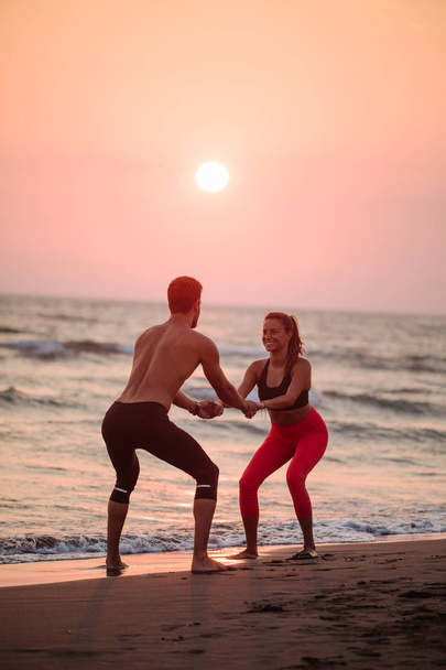 Handsome Caucasian man and beautiful smiling woman doing squats on sandy beach in sunset. - Valokuva, kuva