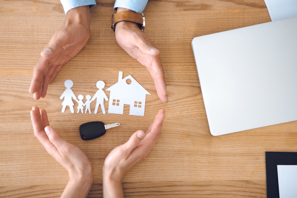 osittainen näkymä vakuutusasiamiesten ja naisten kädet talon, perheen paperimalleja ja auton avain puinen pöytälevy
 - Valokuva, kuva