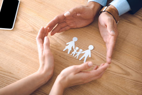 ahşap masa, aile sigortası kavramı üzerinde kısmi görünümü aile kağıt modeli ile eller ve sigorta acenteleri - Fotoğraf, Görsel