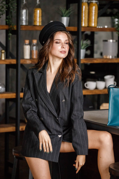 Portrait of beauty female model wearing black jacket and black hat leaning on chair in luxury interior - Valokuva, kuva
