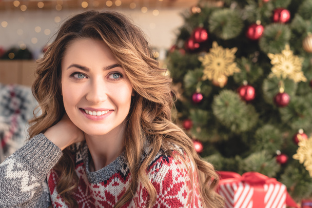 portrait of attractive woman in winter sweater sitting near christmas tree at home and looking away - Foto, Imagem