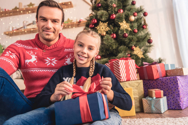 Vater umarmt glückliche Tochter und sie öffnet Geschenk in der Nähe von Weihnachtsbaum zu Hause, Blick in die Kamera - Foto, Bild