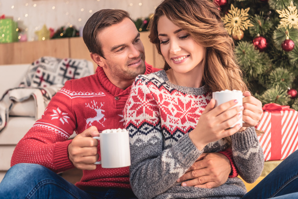 smiling husband and wife holding cups of cappuccino near christmas tree at home - Foto, Imagem