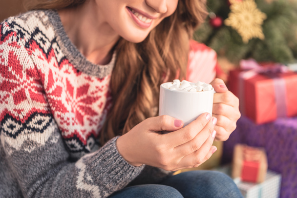自宅クリスマス ツリー近くマシュマロとココアのカップを保持している笑顔の女性の画像をトリミング - 写真・画像
