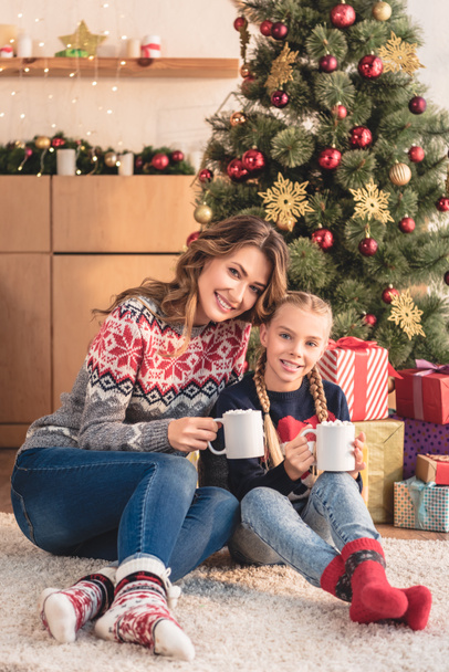 smiling mother and daughter holding cups of cappuccino near christmas tree at home - Φωτογραφία, εικόνα