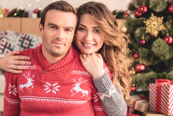 sonriente esposa abrazando marido y mirando a la cámara en casa, árbol de Navidad en el fondo
 - Foto, imagen