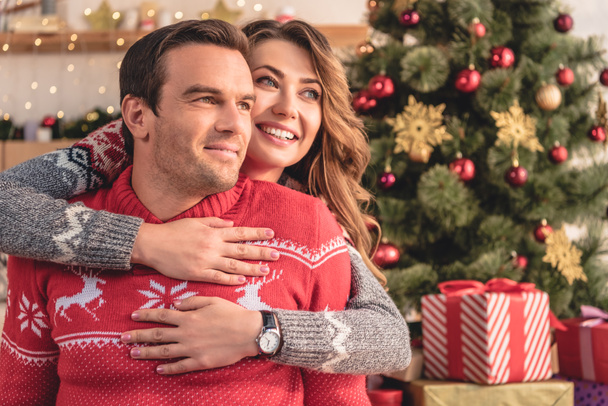 sonriente esposa abrazando marido y mirando hacia fuera en casa, árbol de Navidad en el fondo
 - Foto, imagen
