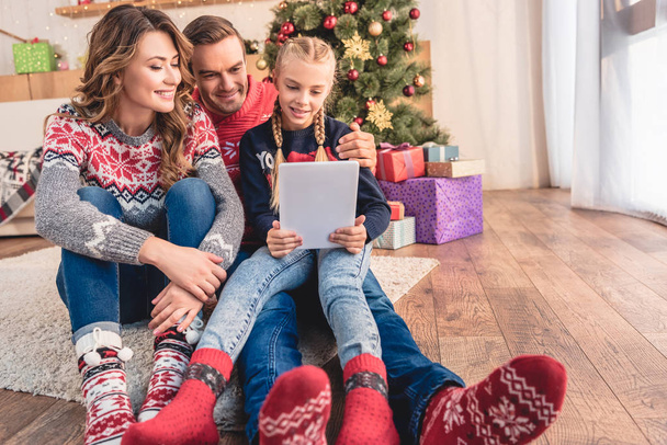 mother, father and daughter using tablet together near christmas tree at home - Фото, изображение