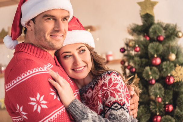 husband and wife in santa hats cuddling near christmas tree at home and looking away - Foto, imagen