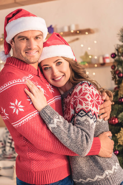 smiling husband and wife in santa hats hugging near christmas tree at home and looking at camera - Fotó, kép