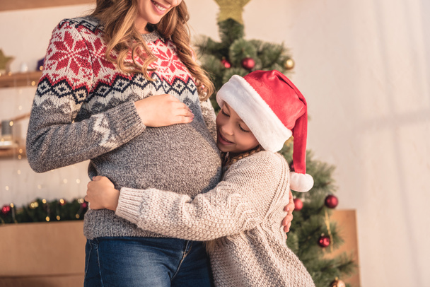 cropped image of daughter in santa hat hugging pregnant mother near christmas tree at home - Foto, Imagem