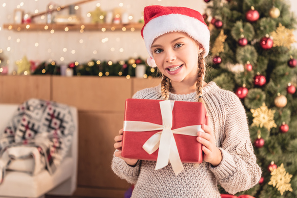 adorable preteen kid in santa hat showing gift box and looking at camera at home - Fotografie, Obrázek