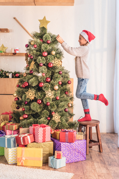 happy child in santa hat standing on stool and decorating christmas tree with gift boxes - Fotó, kép