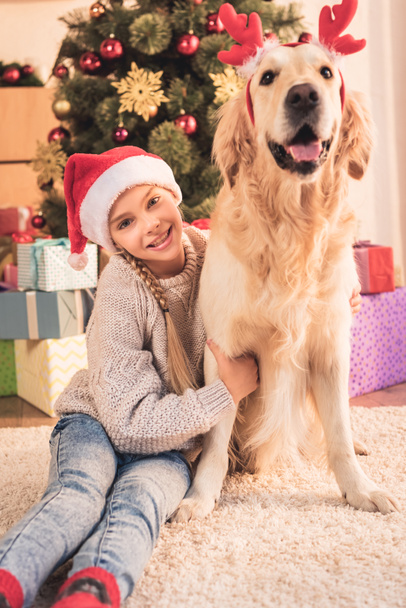 smiling kid in santa hat and golden retriever dog with deer horns sitting near christmas tree with gifts - Photo, Image