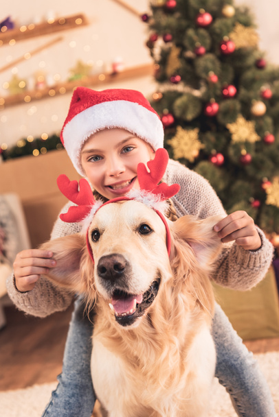 happy child in santa hat and dog with deer horns having fun at home near christmas tree - Photo, Image