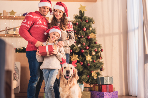 smiling family in santa hats with dog in deer horns standing near christmas tree with gift boxes - Zdjęcie, obraz